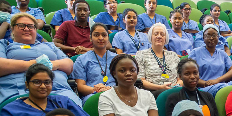 Internationally educated nurses - Asian, predominantly black and two white nurses sitting in an audience cropped to show 18  people mostly wearing a light blue uniform and sitting on green chairs