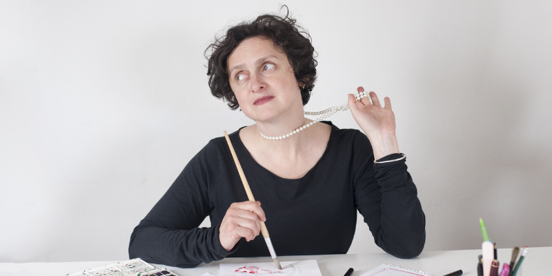 woman sat at a table painting while holding her necklace