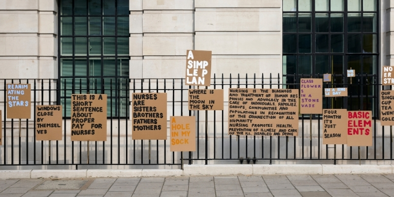 An image of the external black metal fencing which rings around the Royal College of Nursing. Signs of various sizes painted by artist Peter Liversidge have been put up all along it with various quotes and statements on them. From left to right they read 'Reanimating the stars'; 'Windows close by themselves'; 'This is a short sentence about proper pay for nurses'; 'Nurses are sisters brothers fathers mothers'; 'A hole in my sock'; 'A simple plan'; 'Where is the moon in the sky'; 'Nursing is the diagnosis and treatment of human responses and advocacy in the care of individuals, families, groups, communities and populations in recognition of all humanity. Nursing promotes health, prevention of illness and care of the ill, disabled and dying'; 'Glass flowers in a stone vase'; 'Save the NHS'; 'In a single day'; 'Four months it's just a season'; 'We do'; and 'Basic elements'.