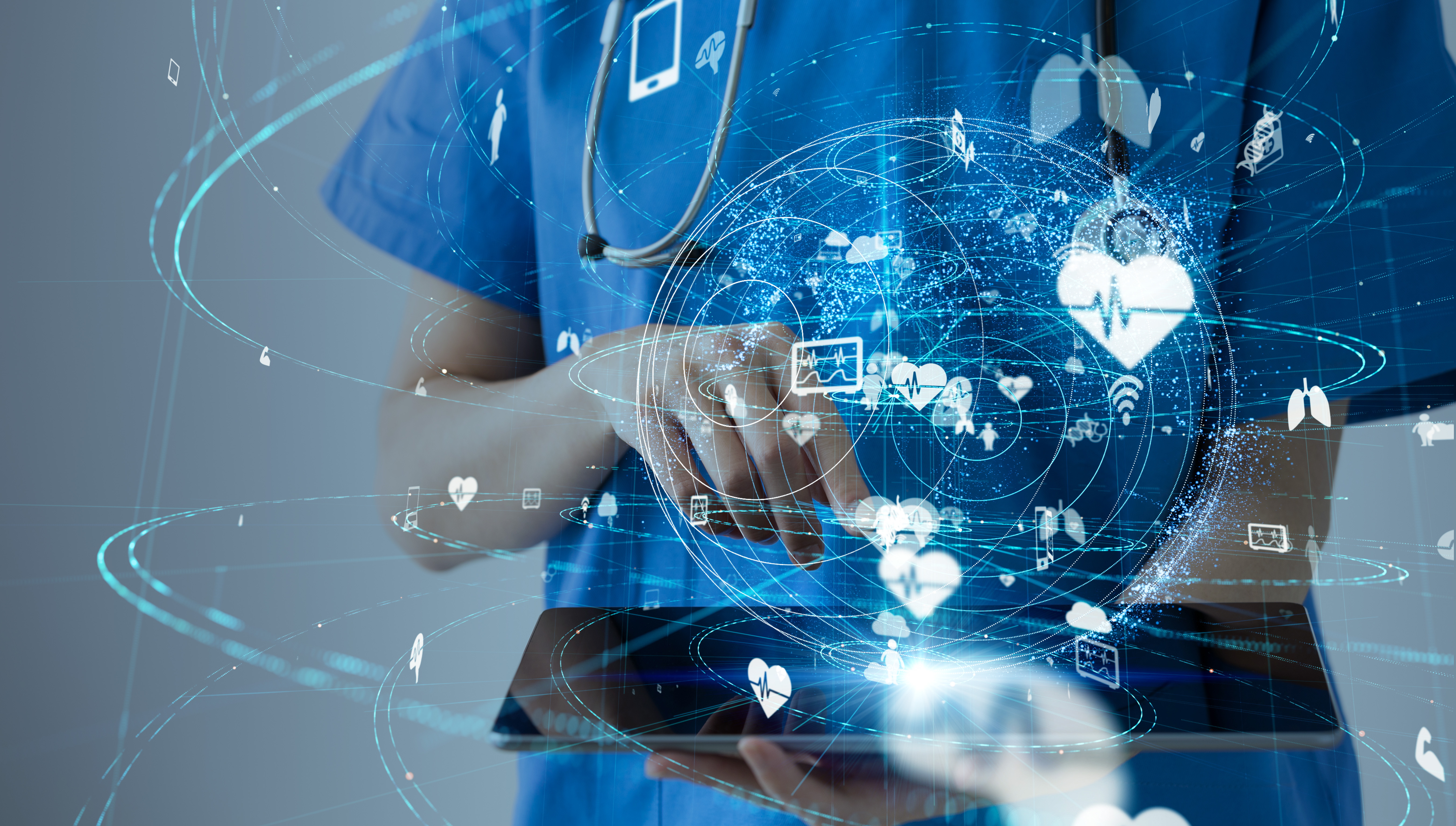 A nurse in blue scrubs interacts with a digital tablet, holding it horizontally in one arm with the other hand poised over the screen. Only the nurse's midriff is visible, but the earpieces of a stethoscope are visible hanging down from the neck. Holographic medical icons, including hearts and lungs, are projected from the device, symbolising innovative health technology.