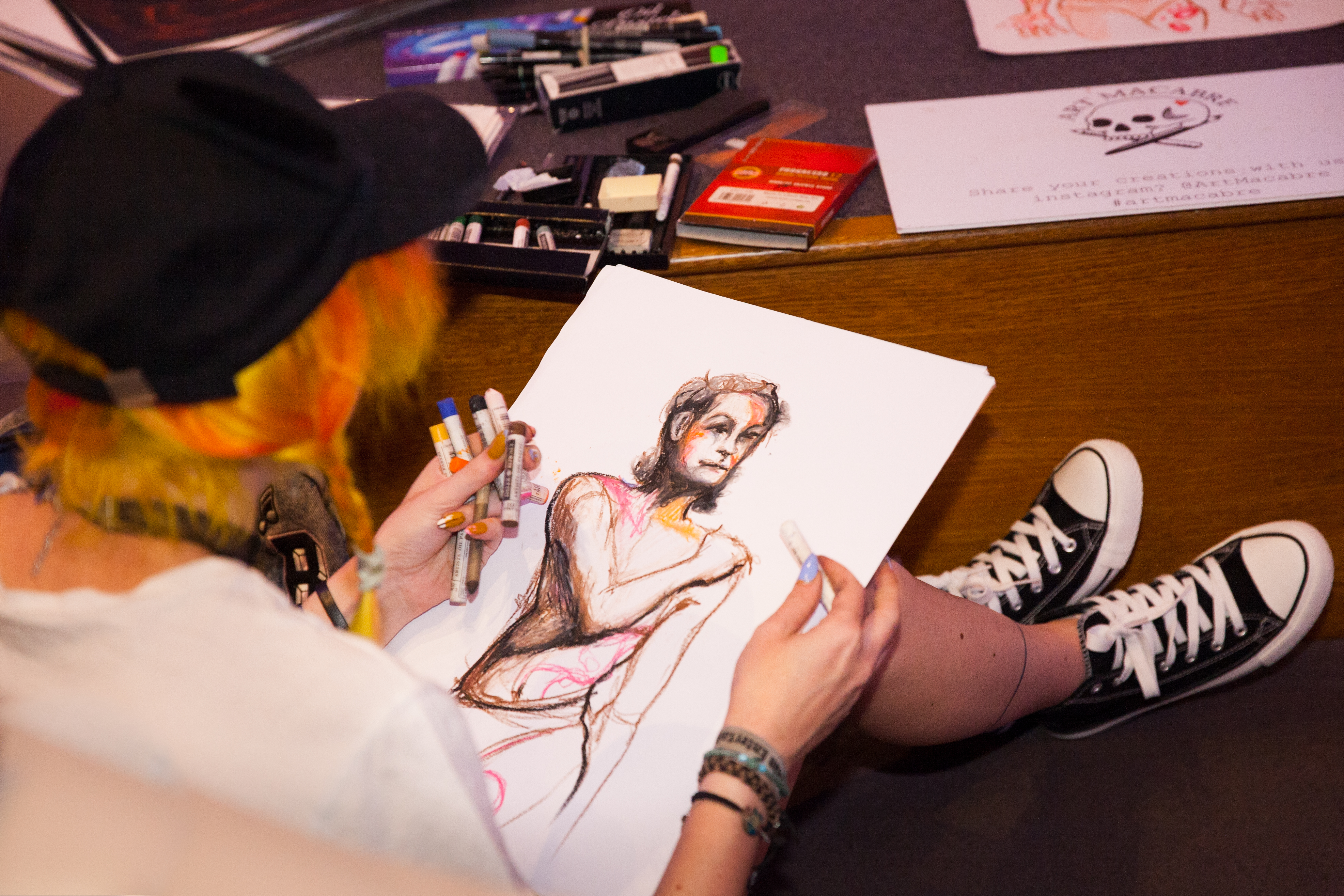 An artist sitting on the floor, drawing a colourful portrait of a person from the torso upwards on A4 paper, surrounded by art supplies. The artist, who has yellow and orange hair in two plaits, has their back to us, and is wearing a black cap and black converse shoes.