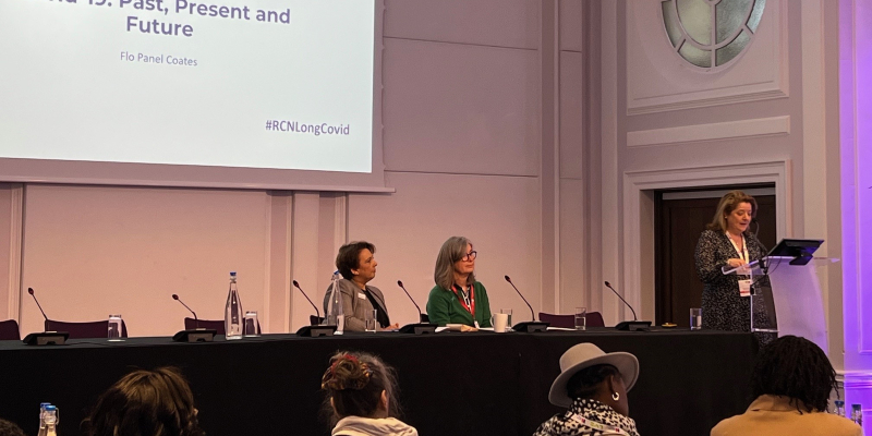 A wide shot is seen, of an event about long COVID held at RCN headquarters. Seen seated are RCN General Secretary and Chief Executive Professor Nicola Ranger and Patricia Marquis, Executive Director of RCN England. Keynote speaker Flo Panel Coates is standing at a lectern giving a speech.