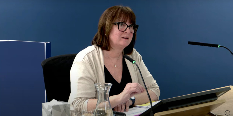 The RCN's Rose Gallagher is seen giving evidence at the COVID inquiry on 18 March 2025. She is wearing a black top with a cream cardigan over the top. She wears black-framed glasses, and sits on a chair with two microphones in front of her. On the table are seen a box of tissues, a jug of water and various papers.