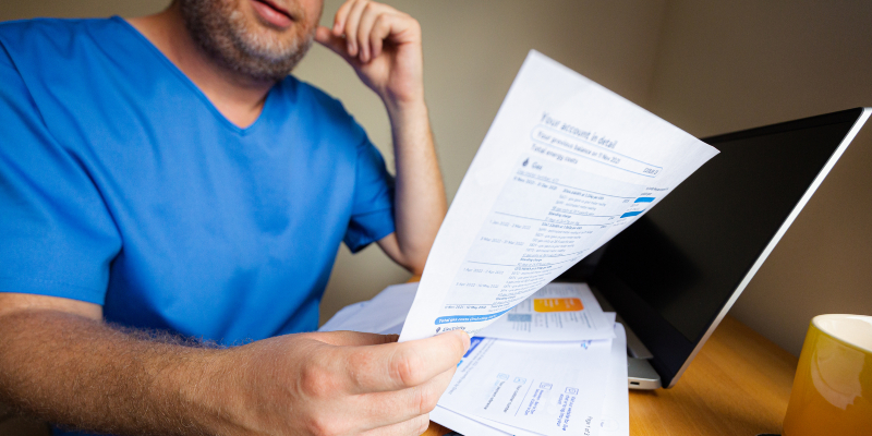 A male nurse in his 30s checks his energy bills at home. He is wearing blue scrubs as he has just come home from work. The man has a worried expression as he examines the paper documents